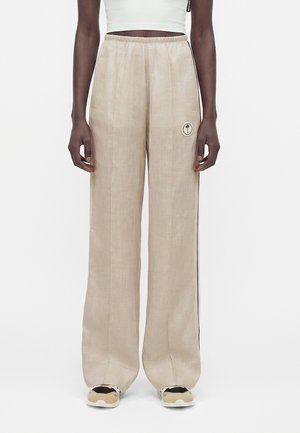 Pantaloni bej cu croială largă și talie elastică, cu un mic logo circular cu palmier pe coapsa stângă, purtați cu adidași bej și negri.