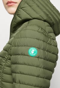 Doudoune verte olive avec un design matelassé horizontal, fermeture éclair et un patch logo rond vert sur l'épaule, présentant un graphique de canard.