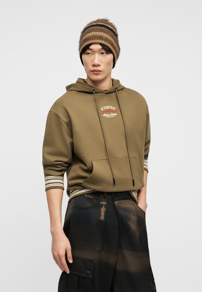 Νεαρό άτομο που φοράει ελιά πράσινο φούτερ με λευκές ρίγες στα μανίκια, καφέ ριγωτό σκουφάκι και μαύρο παντελόνι cargo με πιτσιλιές μπογιάς.