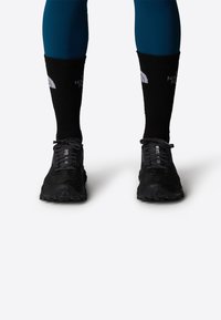Zapatillas de trail running negras con parte superior de malla texturizada y suela de goma. Combinadas con calcetines negros hasta el tobillo que presentan un logo blanco.