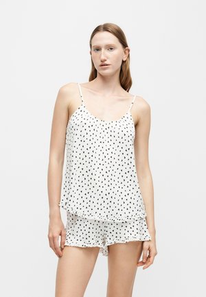 Jeune femme portant un débardeur blanc et un short avec de petits motifs de cœurs noirs sur un fond uni.