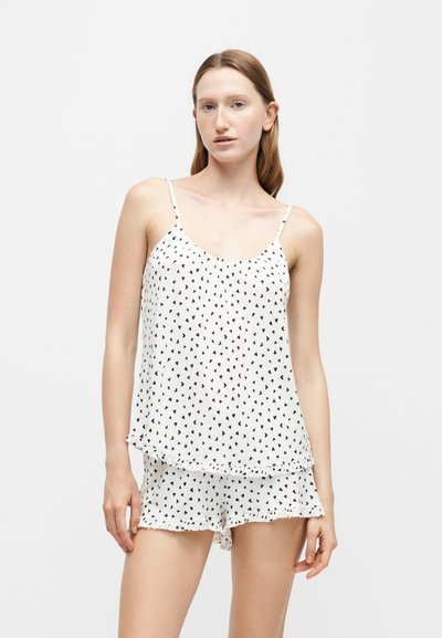 Jeune femme portant un débardeur blanc et un short avec de petits motifs de cœurs noirs sur un fond uni.