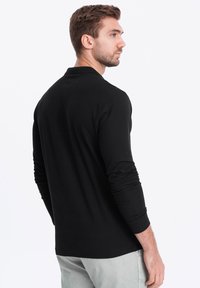 Musta pitkähihaisen poolon paita, jossa on rento istuvuus, pehmeä rakenne ja klassinen kaulus. Takana on sileä pinta ja vähäiset saumat.