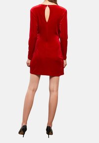 Robe en velours rouge avec des manches longues, une silhouette cintrée et un dos avec un détail en trou de serrure. Associée à des chaussures à talons hauts noires.