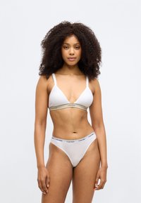 Balta kokvilnas bralette ar trīsstūrveida formu un bēša Calvin Klein zīmolu joslu; saskaņota ar atbilstošām baltām biksītēm, kas rotātas ar melnu logotipa joslu.
