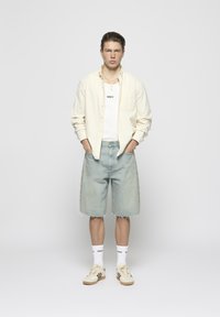 Model wearing a light beige button-up shirt over a white tank top, paired with light wash denim shorts and beige sneakers.