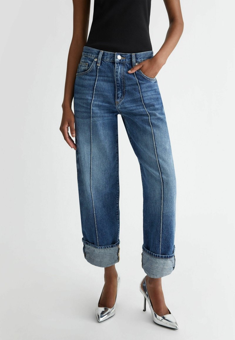 Jeans bleus amples avec revers retroussés, soulignés par des coutures contrastantes, associés à des chaussures à talons argentées. Haut noir ajusté.