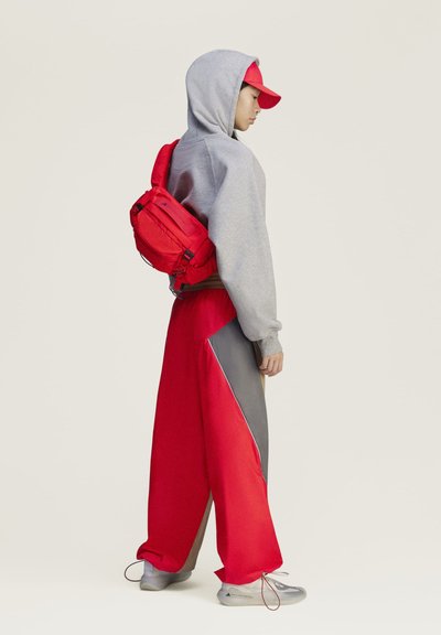 Persona que lleva una sudadera con capucha gris, gorra roja, pantalones anchos rojos y grises, zapatillas blancas y una mochila roja, de perfil sobre un fondo liso.