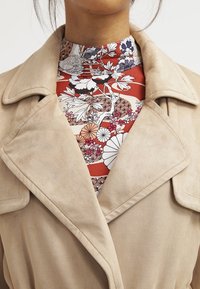 Kvinna som bär en beige mockajacka över en höghalsskjorta med ett flerfärgat blommönster och fåglar på en rostfärgad bakgrund.