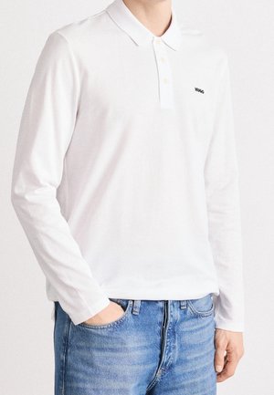 Osoba ubrana w białą długą koszulkę polo z logo "HUGO" na piersi i niebieskie dżinsy, stojąca z lewą ręką w kieszeni.