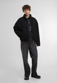 Veste en polaire noire avec un col haut, fermeture éclair, coupe décontractée et poches latérales. Portée sur un t-shirt graphique noir et un jean sombre.