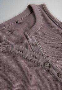 Primer plano de una camisa de tela acanalada color malva con cuello redondo y tapeta de botones con cuatro botones sobre una superficie plana.