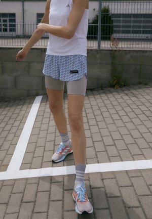 Osoba ubrana w biały tank top, niebieskie wzorzyste spodenki na szarych spodenkach kolarskich, w paskowane skarpetki oraz szaro-niebieskie buty do biegania stojąca na utwardzonej nawierzchni.