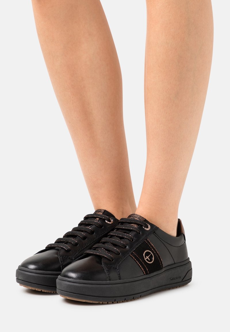 Zapatillas de cuero negro con acentos texturizados, cordones metálicos y una suela de goma gruesa, con un logo en el costado y un diseño de punta redondeada.