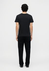 Άνδρας με κυματιστά μαλλιά που φοράει απλό μαύρο κοντομάνικο μπλουζάκι και μαύρο παντελόνι, στέκεται με την πλάτη προς τα εμπρός σε λευκό φόντο.