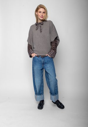 Blonde vrouw staat, draagt een grijze cape trui over een blouse met patroon, opgerolde losse blauwe spijkerbroek en zwarte enkellaarzen tegen een witte achtergrond.