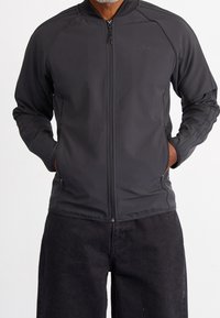 Černá sportovní zipová bunda s stojáčkem, vyrobená z strukturované látky, má boční kapsy na zip a tři pruhy na rukávech.