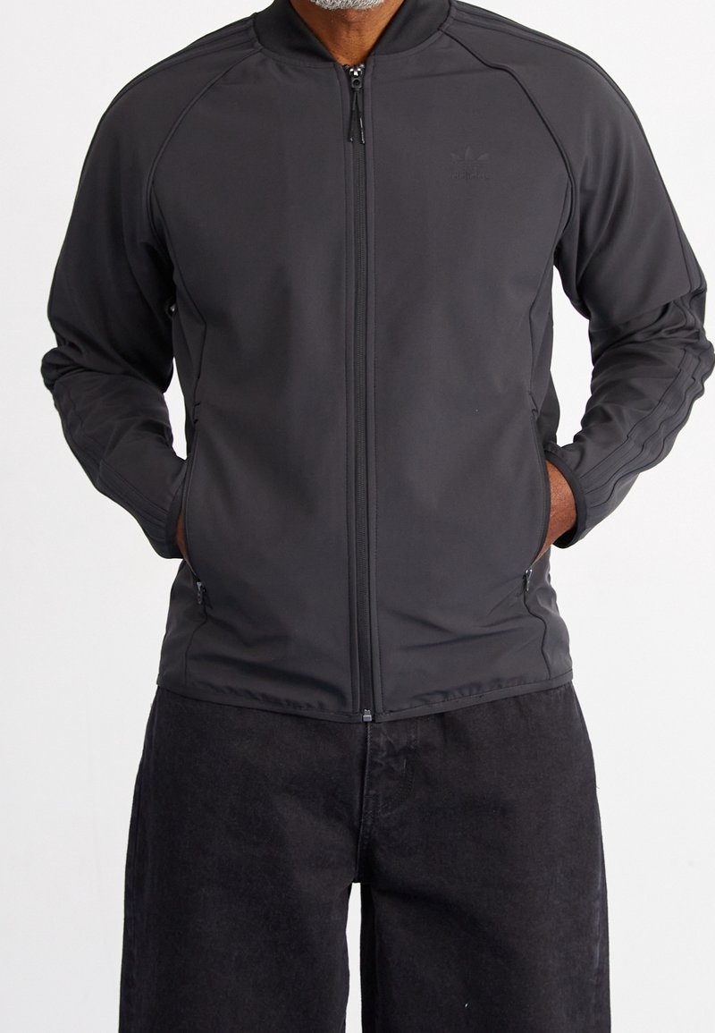 Černá sportovní zipová bunda s stojáčkem, vyrobená z strukturované látky, má boční kapsy na zip a tři pruhy na rukávech.