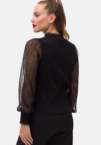 Mustan neulepaidat, joissa on läpinäkyvät, teksturoidut hihat, joissa on herringbone-kuvio ja röyhelömittarit. Klassinen pyöreä pääntie, tiukka muotoilu.