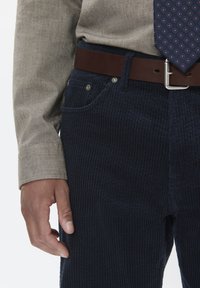 Pantaloni di velluto a coste blu navy con righe verticali, cintura in pelle marrone e camicia beige con maniche a cuffia; cravatta a fantasia visibile. Mano appoggiata vicino alla tasca.