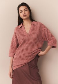 Rožinis megztas megztinis su V formos iškirpte ir trumpomis rankovėmis, tekstūruotas skylutėmis, derinamas su atitinkančia sijonu. Lygus audinys ir laisvas siluetas.