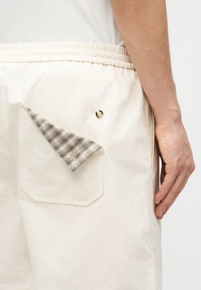 Gros plan sur la main d'une personne à côté d'un short blanc cassé avec une taille élastique, une poche arrière doublée d'un tissu écossais retourné et un petit bouton en détail.