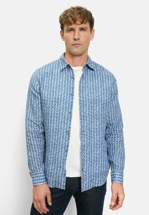 Mann trägt ein aufgeknöpftes blau-weiß kariertes Hemd über einem weißen T-Shirt und dunkle Hosen, steht vor einem einfarbigen hellen Hintergrund.