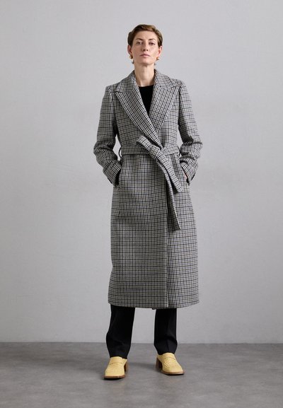 Gray and black plaid long coat with a wrap design, wide collar, and matching belt. Worn over black pants and yellow woven shoes.