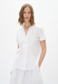 Blusa bianca a maniche corte con scollatura a V e pieghe sottili, realizzata in tessuto leggero con una superficie liscia.