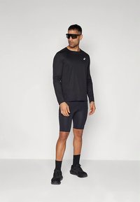 Haut de sport noir à manches longues et short assorti, tous deux de coupe élégante et ajustée, complétés par des chaussures de sport noires et des lunettes de soleil.