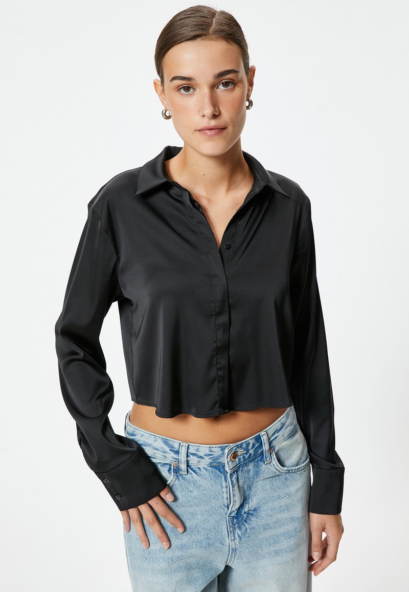 Camisa negra corta de tela suave con botón en el frente, mangas largas y cuello con solapa. Combinada con vaqueros de un azul claro.