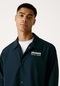 Chaqueta abotonada en color navy, hecha de una tela suave, cuenta con un collar y un logo visible en blanco que dice "RBR RED BULL RACING" en el pecho.
