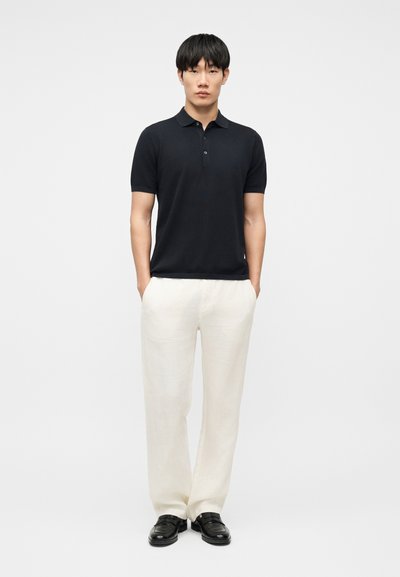 Homme debout les mains dans les poches, portant un polo noir à manches courtes, un pantalon blanc et des chaussures noires, devant un fond uni.