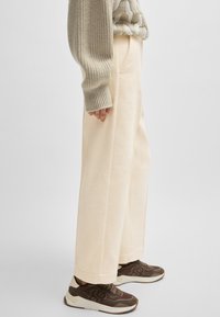 Pantalon à jambes larges beige en tissu texturé avec poches, associé à des baskets marron foncé avec semelle claire et détail logo.