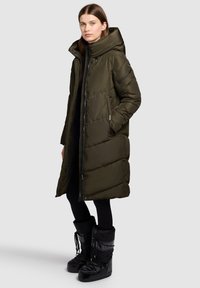 Long olive green puffer coat with a high collar, zip closure, side pockets, and a quilted design. Worn with black snow boots.