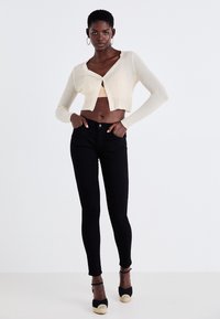 Cárdigan corto de punto acanalado crema con mangas largas, combinado con jeans ajustados negros de tiro alto y sandalias negras de cuña con un acento trenzado.