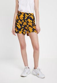 Shorts noirs avec un motif de fleurs jaunes, dotés d'un ourlet festonné. Assortis à un t-shirt blanc et des baskets blanches.