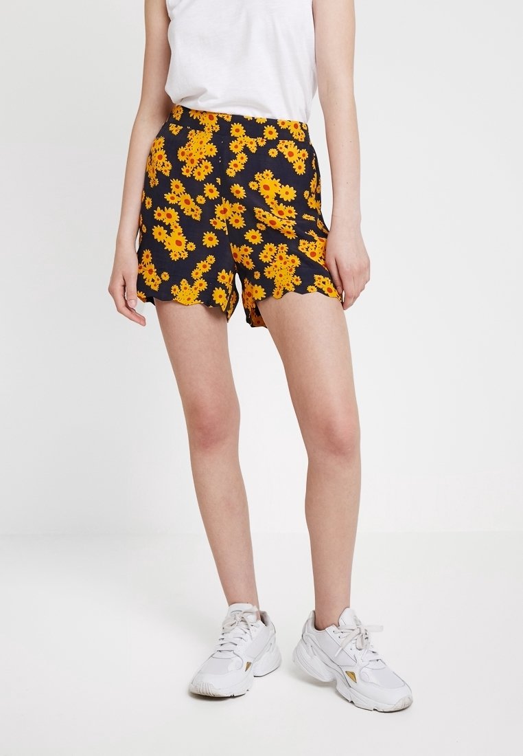 Shorts noirs avec un motif de fleurs jaunes, dotés d'un ourlet festonné. Assortis à un t-shirt blanc et des baskets blanches.