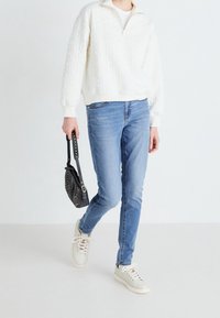 Suéter blanco acolchado con media cremallera, jeans azul claro y zapatillas blancas rotas. Sosteniendo un bolso negro con acentos de tachuelas.