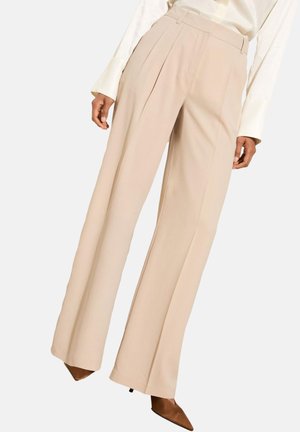 Beige wijde broek met voorplooien, gedragen met een wit blouse met lange mouwen en bruine pumps met spitse neus.