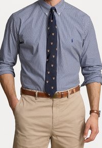 Mężczyzna w niebieskiej i białej koszuli w kratę, z granatowym krawatem w drobne wzory, beżowych spodniach, brązowym pasku i zegarku na nadgarstku.