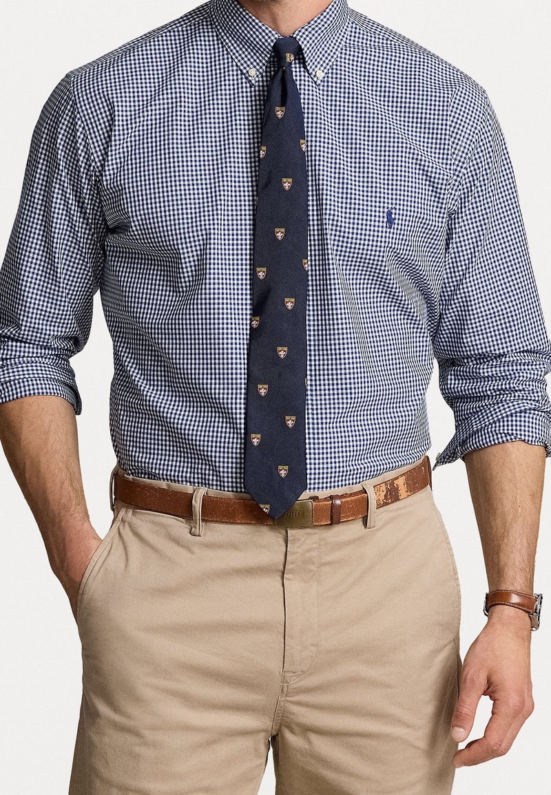 Mężczyzna w niebieskiej i białej koszuli w kratę, z granatowym krawatem w drobne wzory, beżowych spodniach, brązowym pasku i zegarku na nadgarstku.