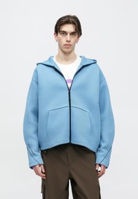 Φωτεινή γαλάζια φούτερ με φερμουάρ, κατασκευασμένη από μαλακό ύφασμα, με μπροστινή τσέπη, άνετη εφαρμογή και επενδεδυμένο κουκούλα. Συνδυασμένη με σκούρο παντελόνι cargo.