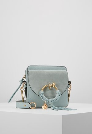 Light blue leather crossbody bag with suede flap, gold circular ring, braided detail, tassels, and a small heart charm on a white surface.