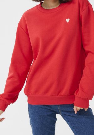 Femme portant un sweat-shirt rouge avec un petit cœur blanc sur la poitrine et un jean bleu, debout les bras légèrement fléchis.