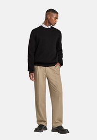 Schwarzer Strickpullover, beigefarbene plissierte Hose, schwarze Schnürschuhe. Das Model steht mit einer Hand in der Tasche vor einem neutralen Hintergrund.