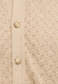 Μπεζ πλεκτό ύφασμα με υφασμένο γεωμετρικό σχέδιο και κάθετη ριμπ ύφανση με δύο διαφανείς κουμπιά με τέσσερις τρύπες.