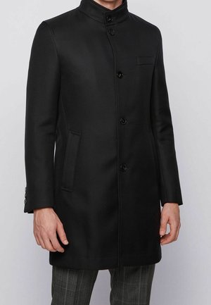 Mężczyzna ubrany w czarny, sięgający kolan płaszcz na guziki z kołnierzem typu stójka oraz w kratkę spodnie, stojący na tle jednolitego szarego tła.