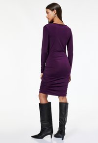 Donna in un vestito lungo a maniche con arricciatura viola aderente, abbinato a stivali neri con tacco al ginocchio, in piedi contro uno sfondo bianco.