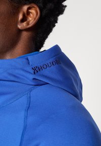 Sudadera azul con un cuello alto, hecha de una tela suave. Presenta el logo "HOUDINI" bordado en azul oscuro cerca de la costura del cuello.